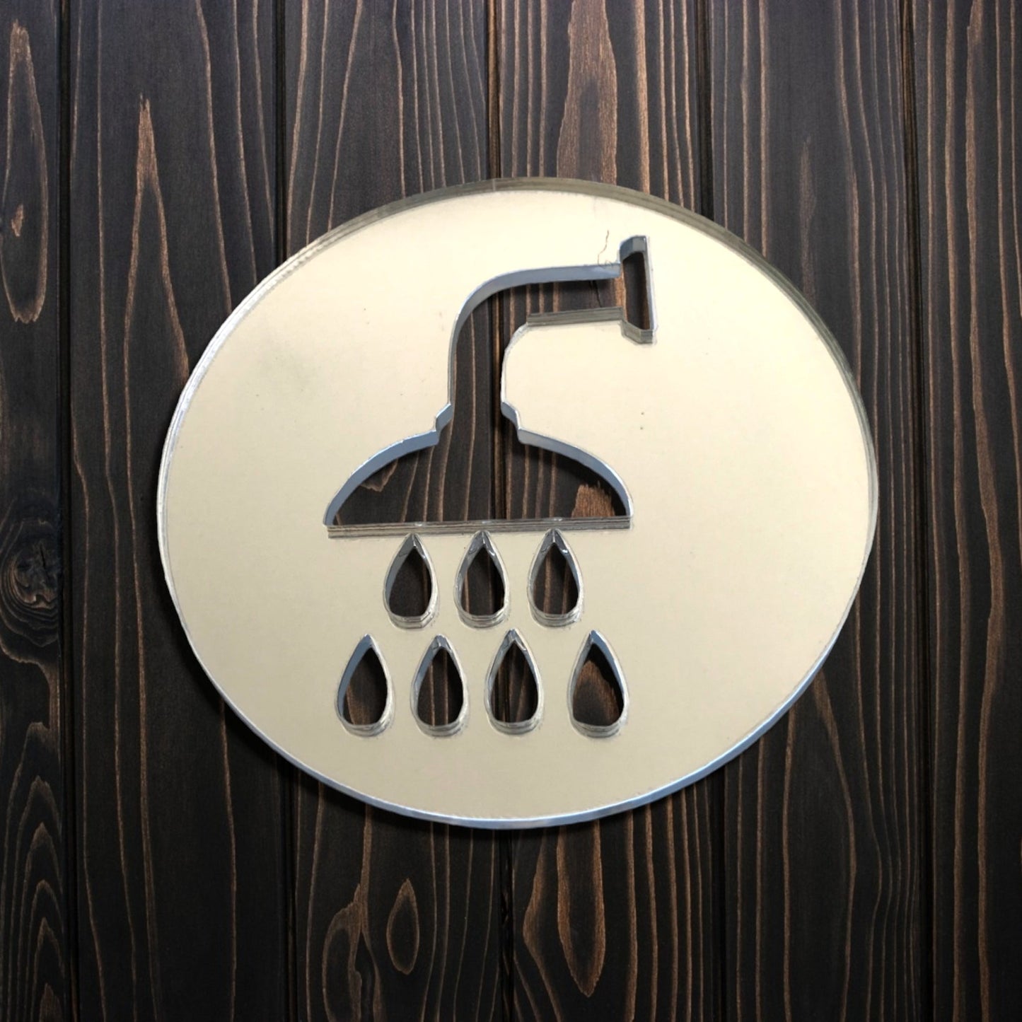 Round mirrored acrylic shower room sign with cutouts of a showerhead and water drops on a dark wood wall.