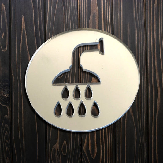 Round mirrored acrylic shower room sign with cutouts of a showerhead and water drops on a dark wood wall.