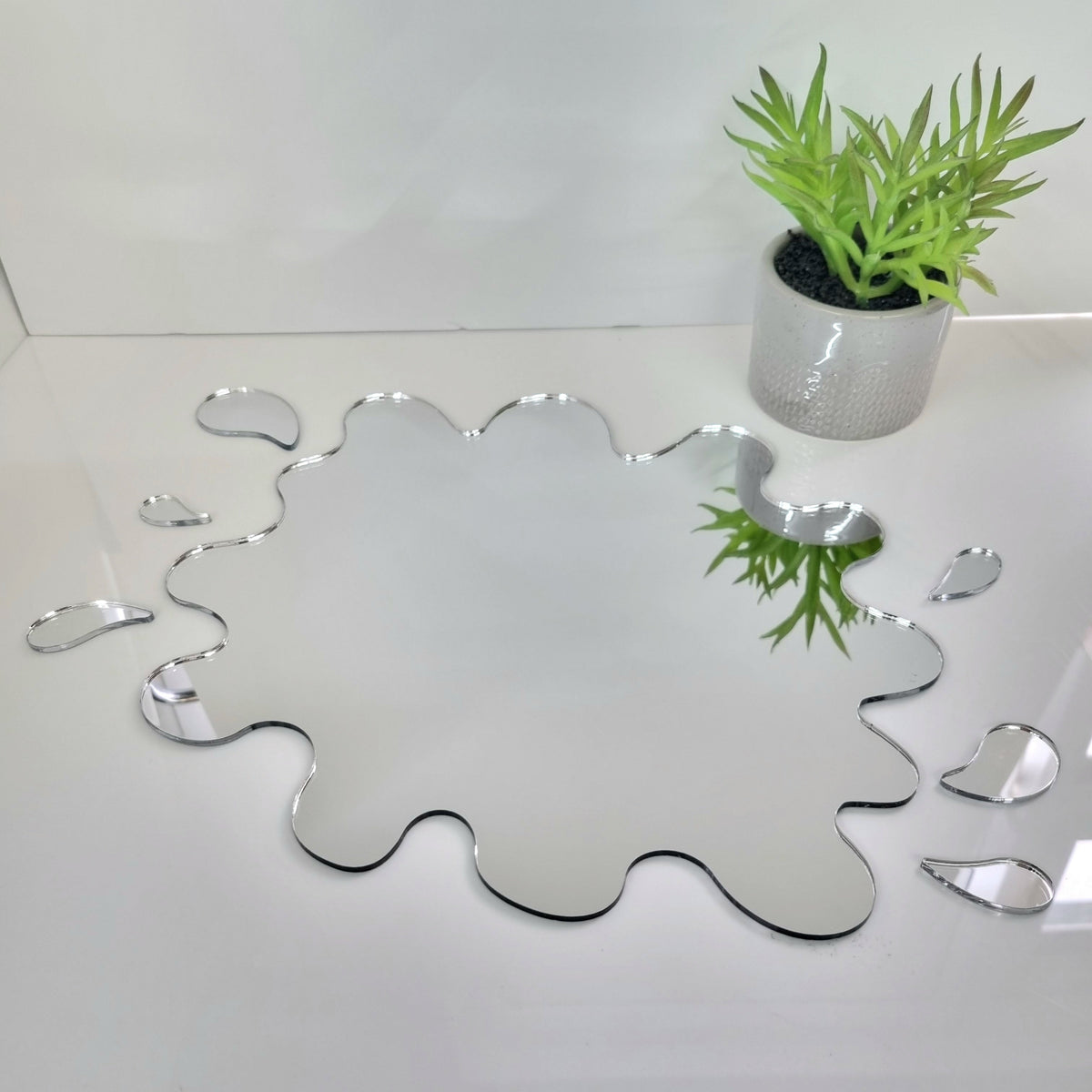 Puddle and six splash mirrors in irregular, organic liquid shapes on a white surface, with a small potted plant nearby.