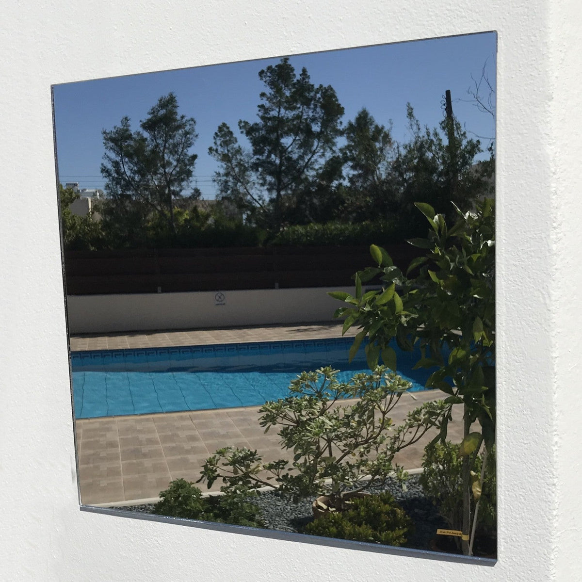 Square garden mirror mounted on a white wall, reflecting a swimming pool, plants, and trees.