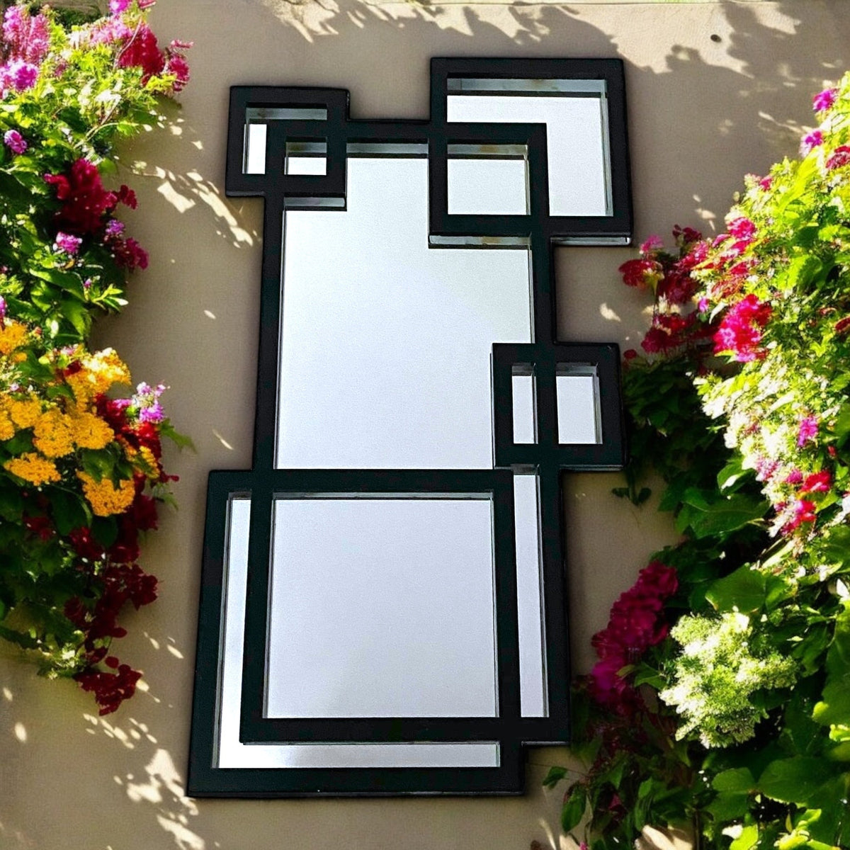 Contemporary black square-framed garden mirror mounted on a wall between blooming flowers.