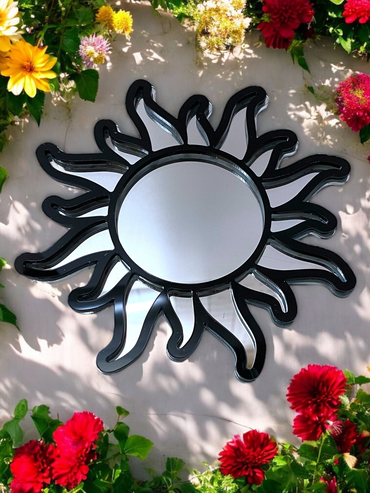 Round sun-framed garden mirror with a black-and-mirror abstract ray design, surrounded by colorful flowers.