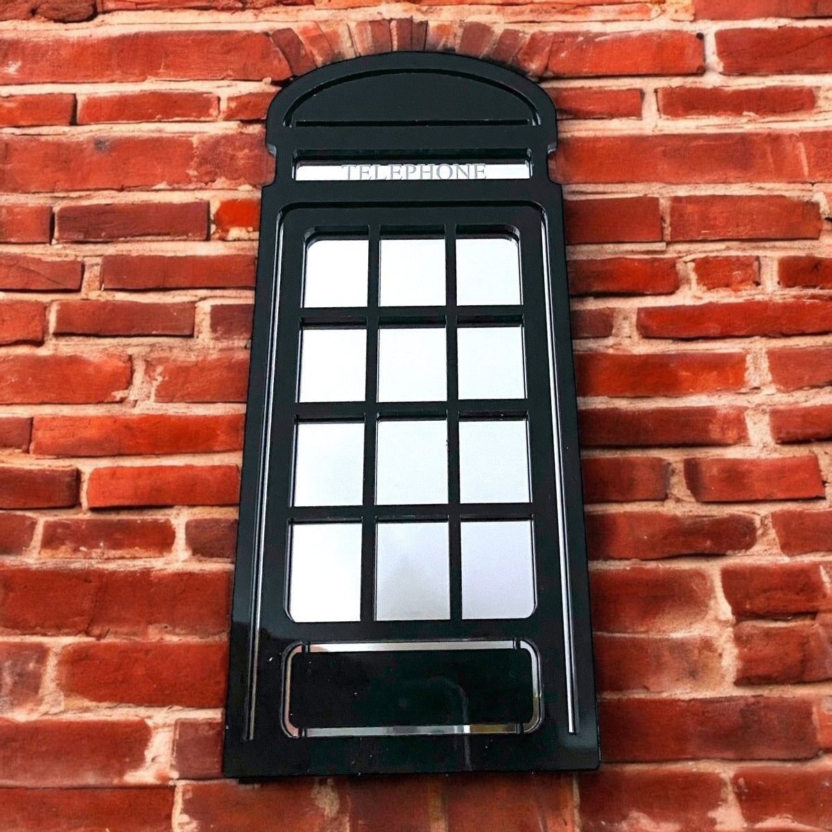 Black British telephone box-shaped garden mirror mounted on a red brick wall.