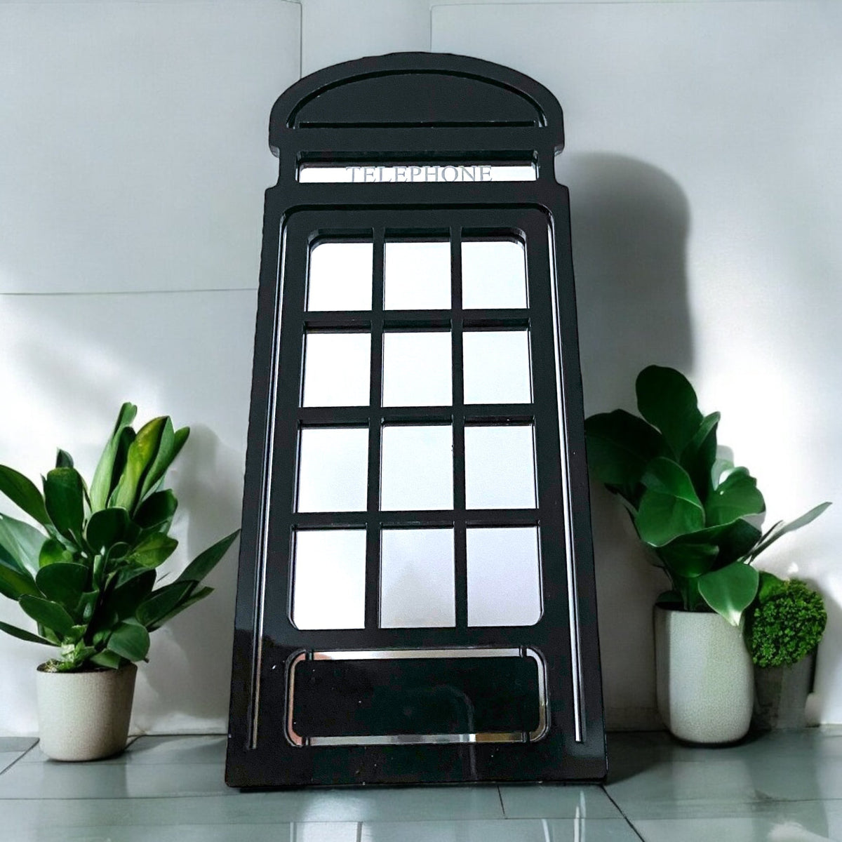 Black wooden telephone box-shaped garden mirror flanked by potted green plants on a grey tiled surface.