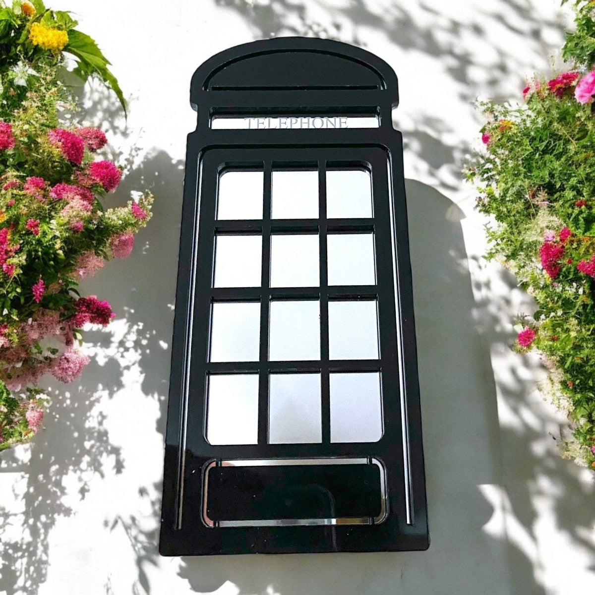 Black British telephone box garden mirror on a white wall between flowers.