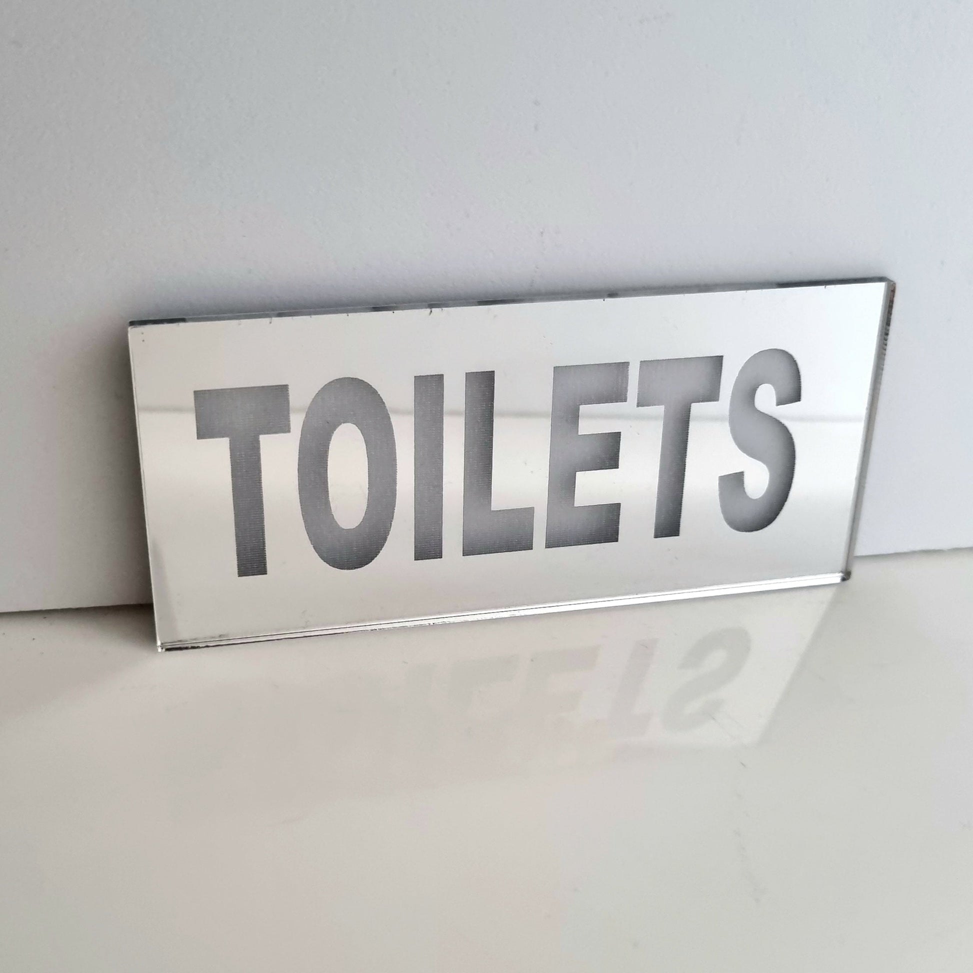 Rectangular mirrored sign with the word "TOILETS" in bold uppercase letters on a white surface.
