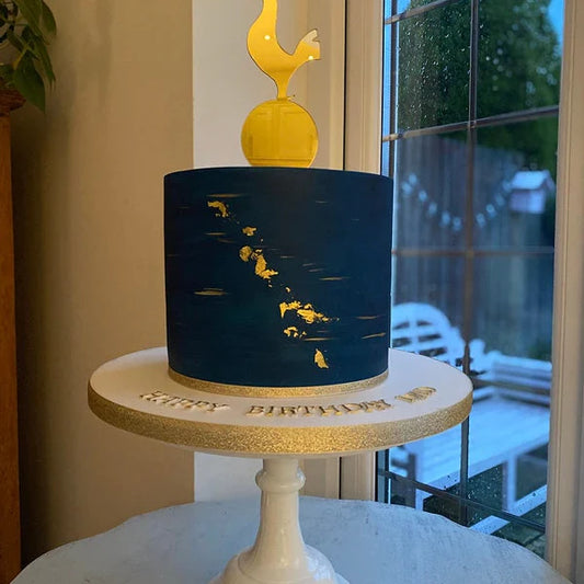 Gold acrylic THFC cockerel cake topper on a navy cake with gold leaf accents and "HAPPY BIRTHDAY" on the board.