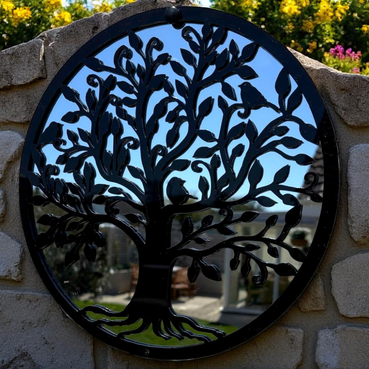 Round black metal garden mirror featuring a Tree of Life motif with branches, leaves, and birds, mounted on a stone wall.