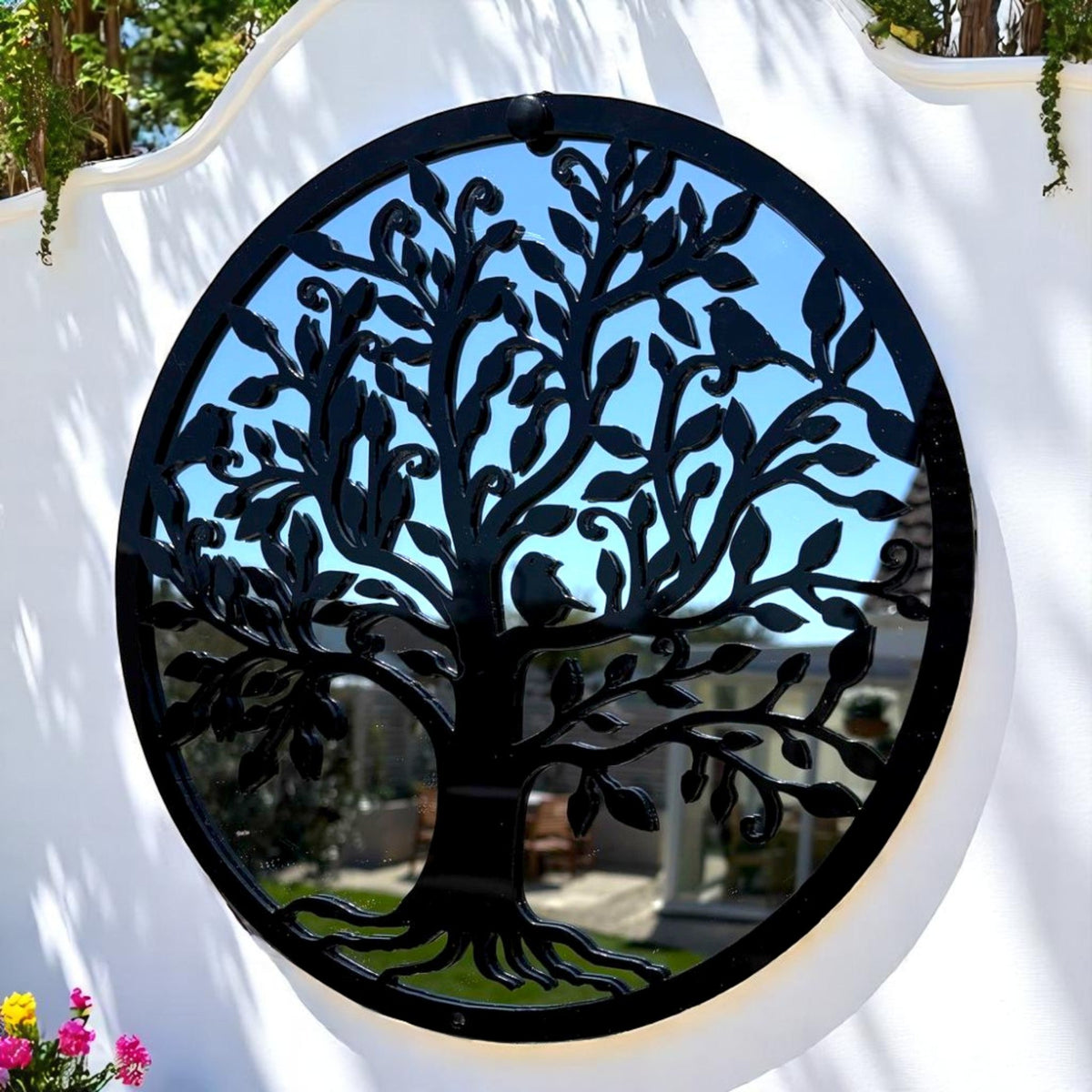 Round black metal Tree of Life garden mirror mounted on a white wall outdoors.