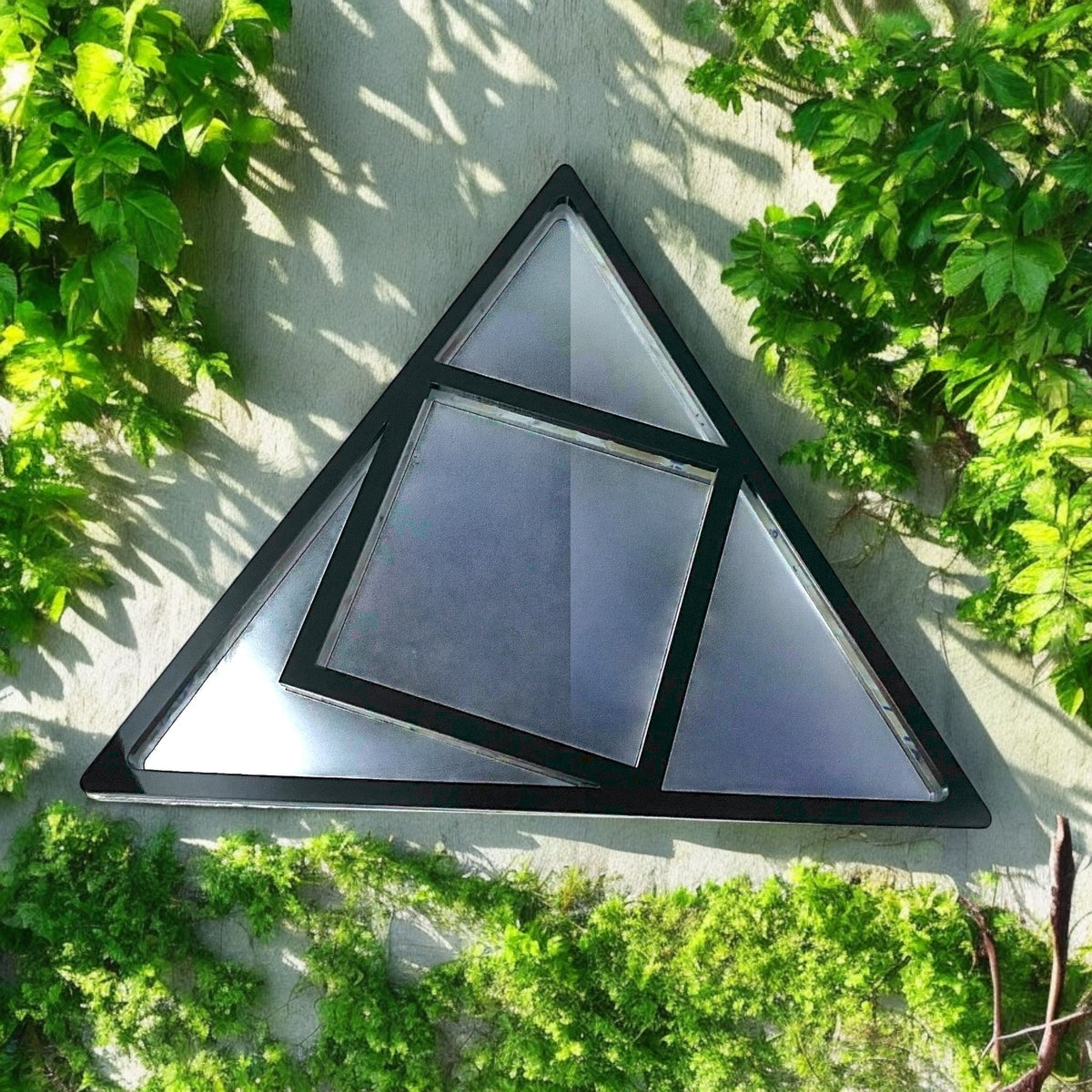 A black-framed triangular garden mirror with an inner square shape, mounted on a green-vined wall.