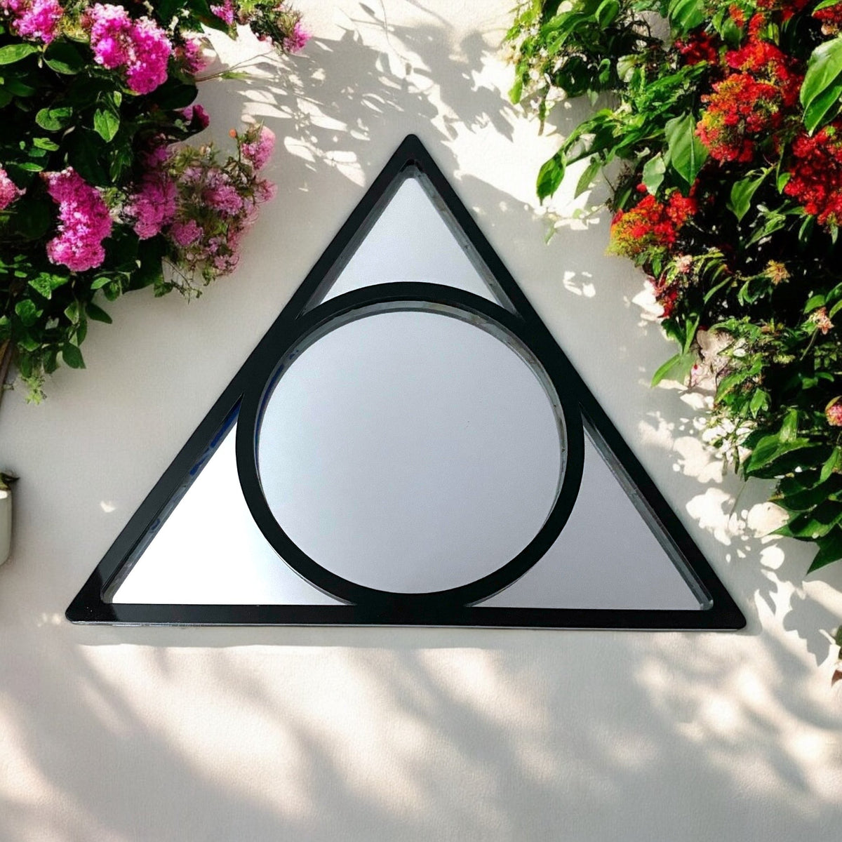 Triangle and circle black metal garden mirror mounted on a white wall between pink and red flowers.