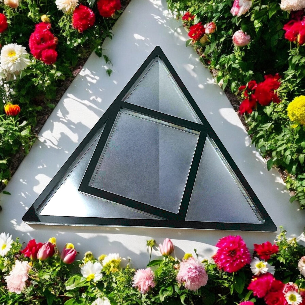 A black metal triangle and square framed garden mirror surrounded by colorful flowers.