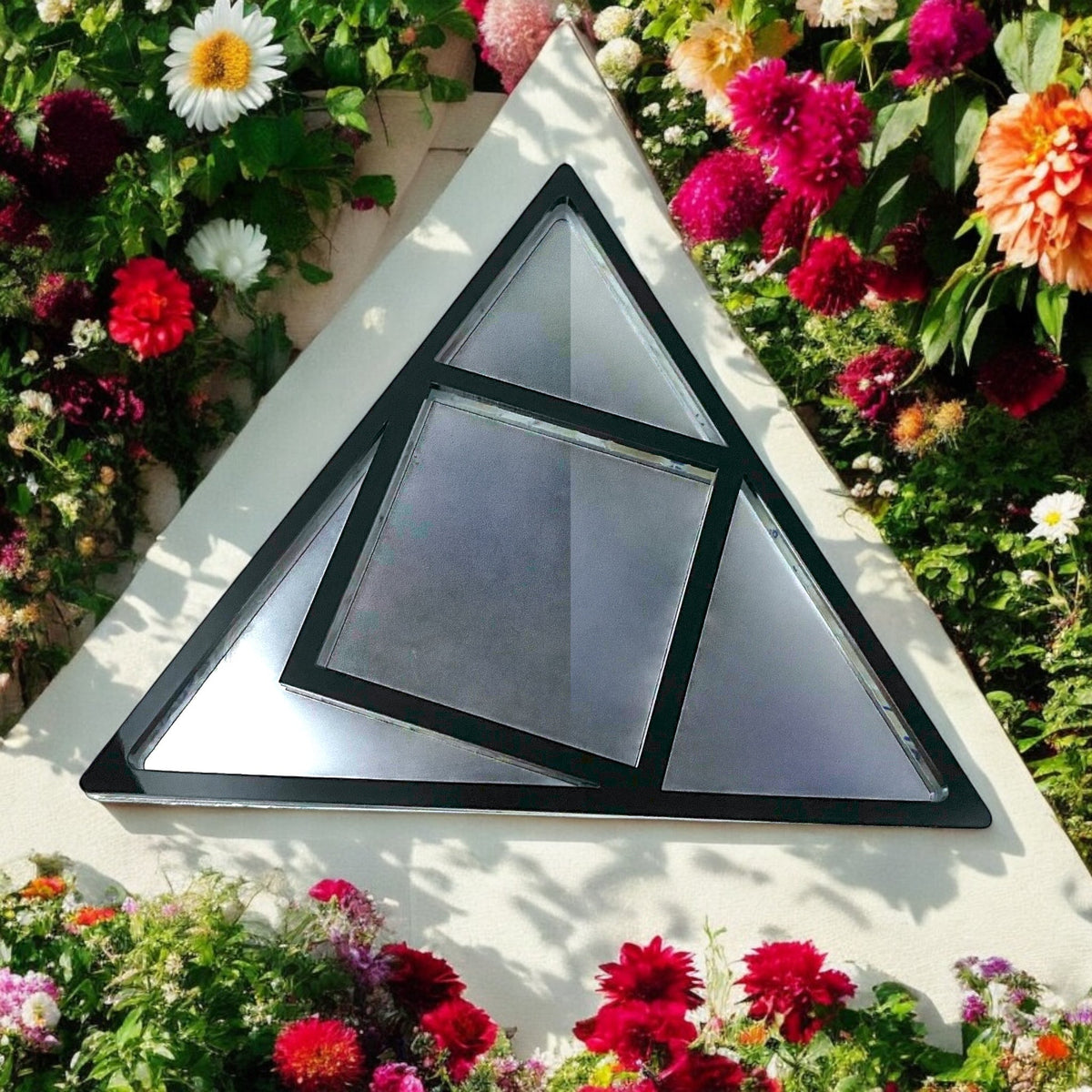 Black-framed triangle garden mirror with intersecting square panes, surrounded by flowers.