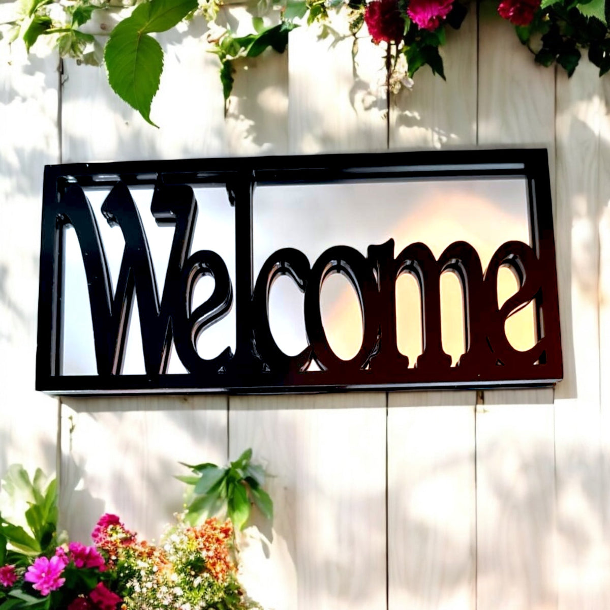 Rectangular black framed garden mirror with cutout "Welcome" lettering hanging on a wooden fence.
