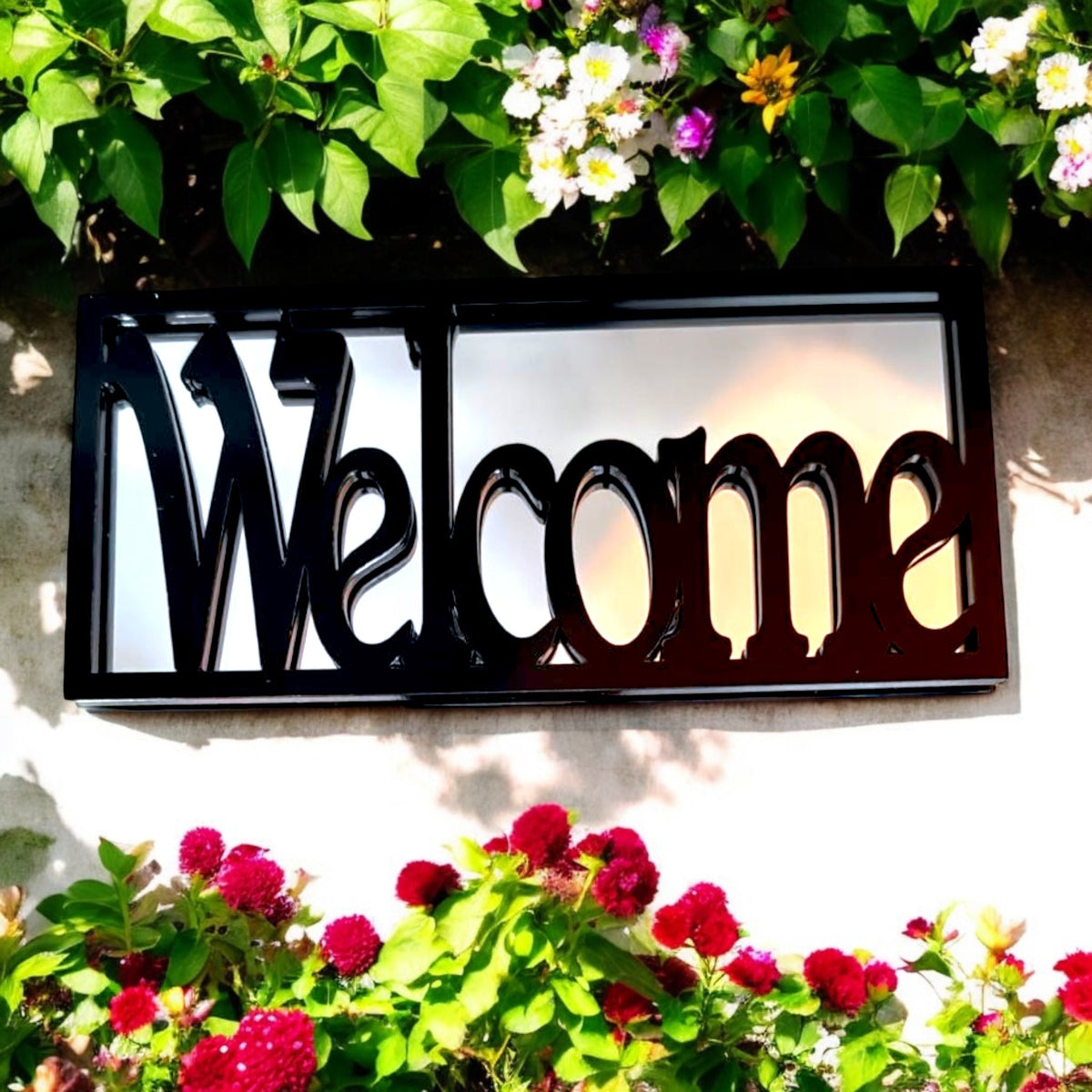 Black framed rectangular garden mirror with the word 'Welcome' overlaid, surrounded by greenery and flowers.