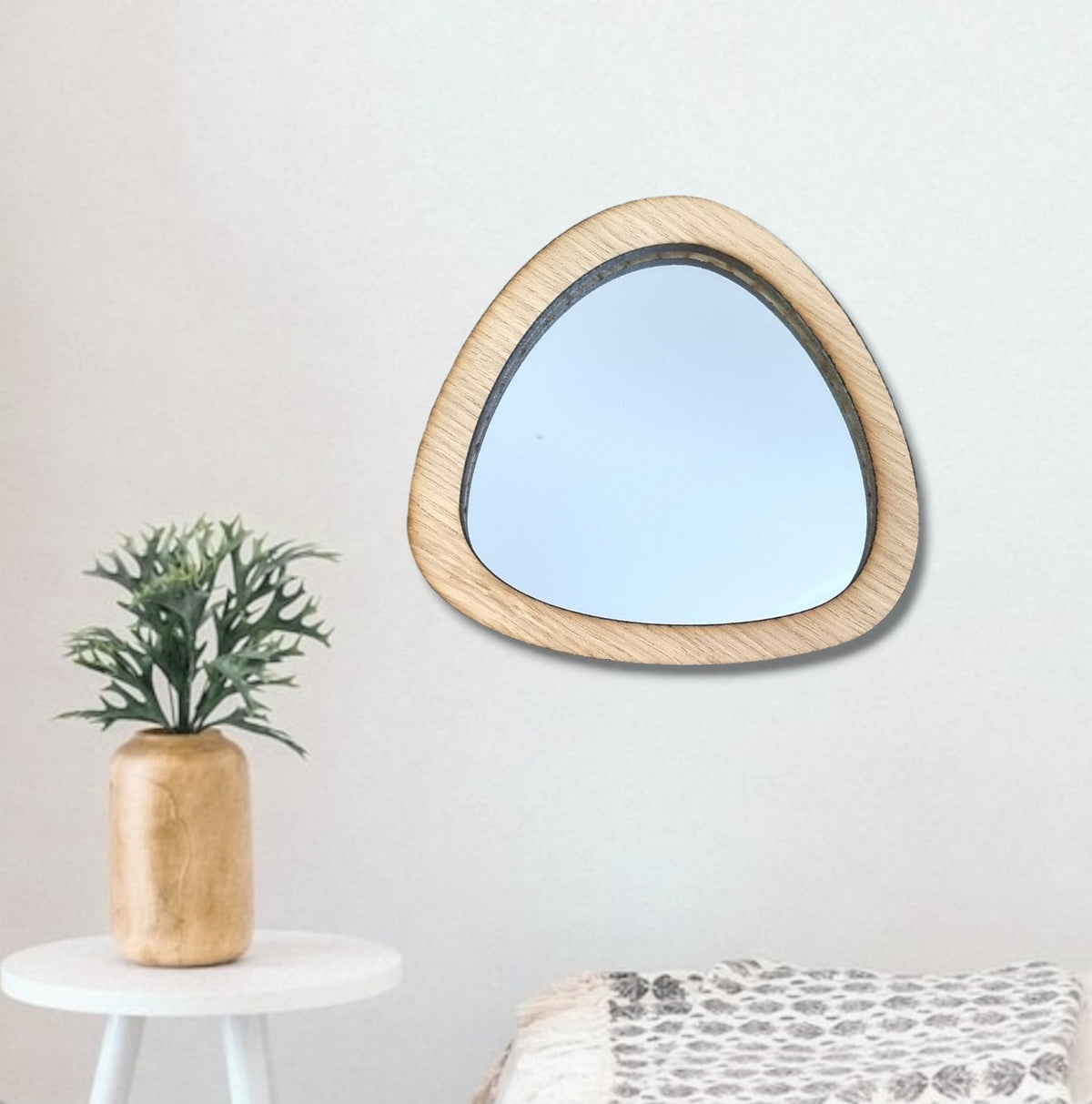 An asymmetric pebble-shaped wall mirror with a light wood frame hanging on a white wall.